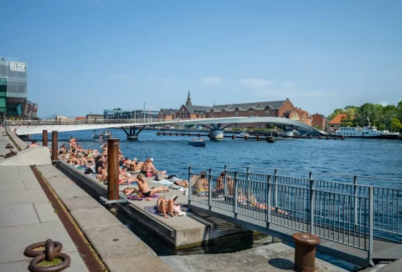 Folk, der solbader på en betonplatform ved en flod, med en moderne fodgængerbro og historiske bygninger i baggrunden under en klar, blå himmel.