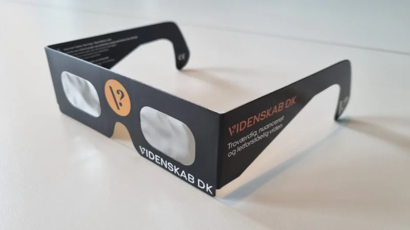 Et par sorte 3D-briller af papir med gennemskinnelige glas og påskriften »VIDENSKAB DK« på stellet, der ligger på en hvid overflade.