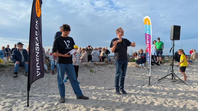 To mænd står på en sandstrand tæt ved nogle flag og en højttaler og taler til en siddende menneskemængde. Den ene mand holder nogle papirer, mens den anden gestikulerer med hænderne. Børn og voksne følger begivenheden under en delvis overskyet himmel.