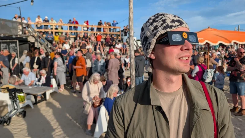 En mand med solbriller og en mønstret hat står på en sandstrand, mens en skare mennesker har samlet sig bag ham under den solrige himmel ved et udendørsarrangement.