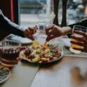 To personer sidder ved et bord på en restaurant, hvor de deler en tallerken med nachos med masser af tilbehør og holder drinks i hånden. Deres hænder rækker ud efter maden, og bordet er dækket med tallerkener, drinks og krydderier.