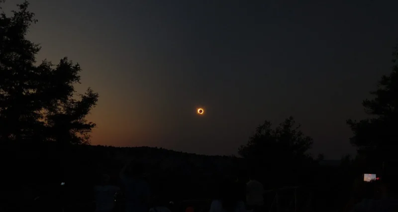 Folk betragter en total solformørkelse ved solnedgang, hvor solens korona lyser klart på den mørke himmel, omgivet af træernes silhuetter og et publikum, der foreviger øjeblikket med kameraer og mobiltelefoner.