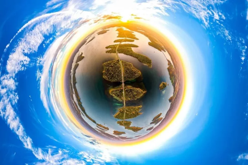 Et 360-graders luftfoto skaber en »tiny planet«-effekt, der viser øer, vand og en vej under en strålende blå himmel med skyer og en gul-orange horisont ved solopgang eller solnedgang.