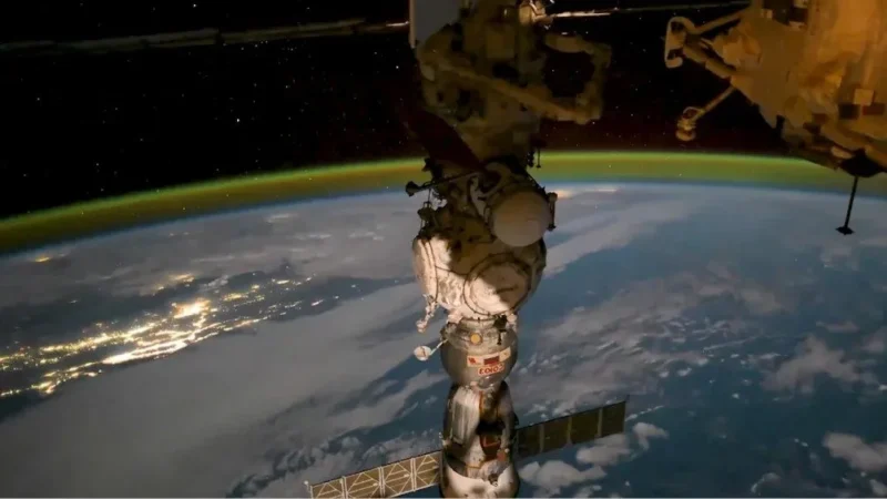 Et rumfartøj, der er koblet til Den Internationale Rumstation, kredser om Jorden, mens byens lys kan ses nedenunder, og et tyndt, glødende atmosfærisk lag strækker sig langs planetens krumning.