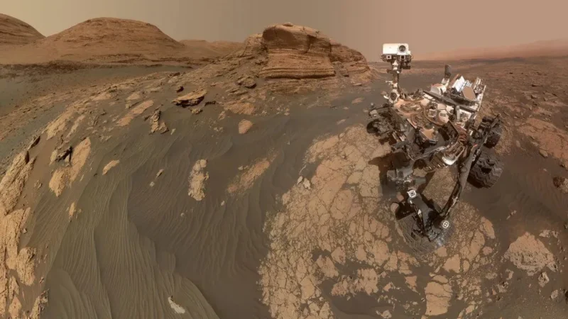 NASAs Curiosity-rover står på et klippe- og sandterræn på Mars med lagdelte bakker og en diset orange himmel i baggrunden. Roverens kamerararm kan ses og fanger en del af sig selv i forgrunden.