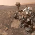 En Mars-robot med kameraer og videnskabelige instrumenter står på en stenet, rødlig Marsoverflade under en diset himmel, omgivet af fjerne bakker.