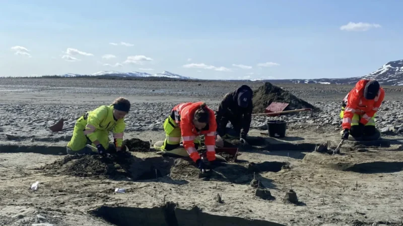 Tre personer i synligt arbejdstøj udfører gravearbejde på en sandet grund udendørs, med bjerge og sne i det fjerne i baggrunden under en klar himmel.
