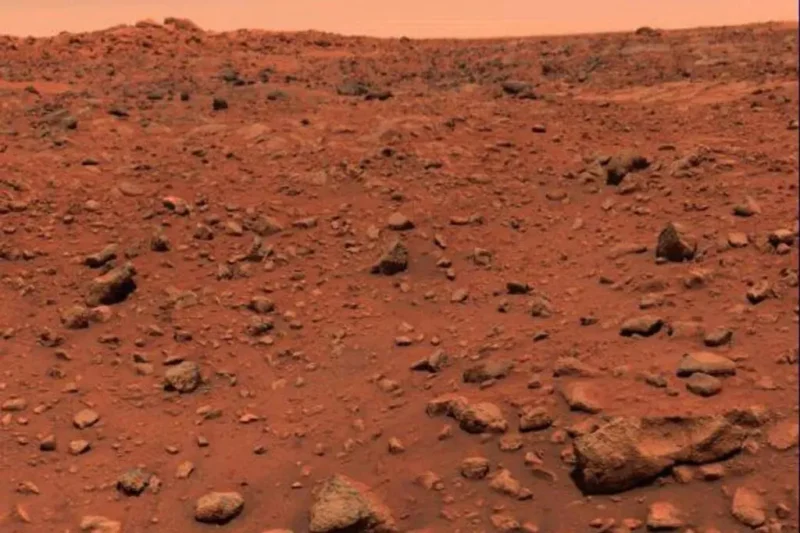 Et klippefyldt, rødligt landskab på Mars, dækket af spredte sten og støv under en diset, orange himmel. Terrænet fremstår goldt og ujævnt og strækker sig mod horisonten uden synlig vegetation eller bygninger.