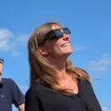 To personer, der bærer beskyttelsesbriller til solformørkelsen, kigger smilende op mod himlen under en klarblå himmel med spredte skyer – et ægte øjeblik af hverdagsvidenskab, hvor nysgerrighed møder forundring.
