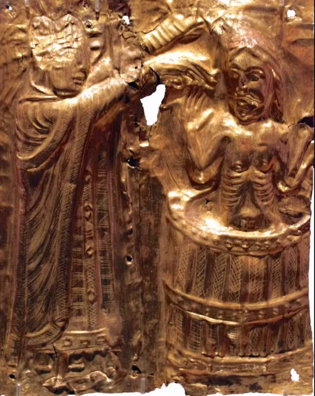 Relief i metal i middelalderlig stil, der forestiller en figur i kappe, der hælder væske over en person, der står i en tønde. Personen i tønden ser ud til at være i nød; tønden og personens klæder er prydet med indviklede mønstre. Nogle områder er slidte eller beskadigede.