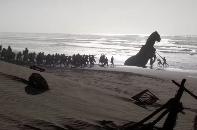En gruppe mennesker trækker en stor, delvist nedgravet statue langs en strand ved solnedgang; bølgerne bruser tæt ved, og der ligger affald spredt ud over sandet.