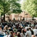 En stor menneskemængde sidder på græsset under træerne og ser en optræden på en udendørs scene med telte og belysning på en solrig dag ved en festival eller et lokalt arrangement.