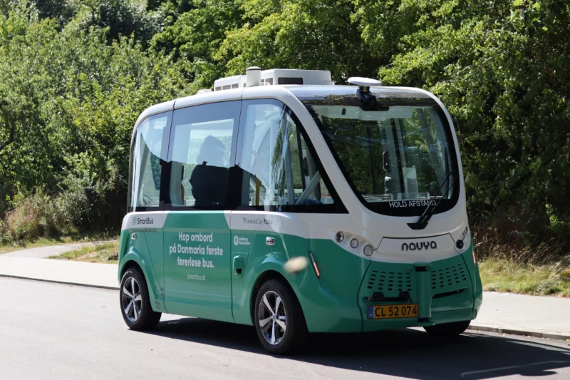 En lille, grøn og hvid selvkørende shuttlebus med dansk tekst på siden holder parkeret ved en vej, med grønne omgivelser i baggrunden. Bussen har ingen synlig chauffør.