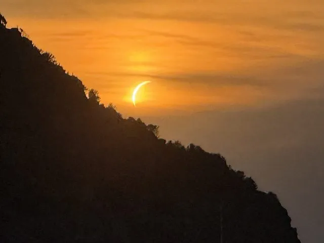 En halvmåneformet sol skjules delvist af et bjergs silhuet ved solnedgang og oplyser himlen med varme orange og gule nuancer.