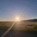 Solnedgang over en græsmark med lange skygger, klar himmel og en række fjerne bygninger, der tegner sig som silhuetter på højre side af horisonten.