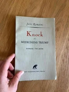En hånd holder bogen *Knock eller Medicinens Triumf* af Jules Romains på en træoverflade. Omslaget er enkelt med et minimalistisk design og sort tekst.