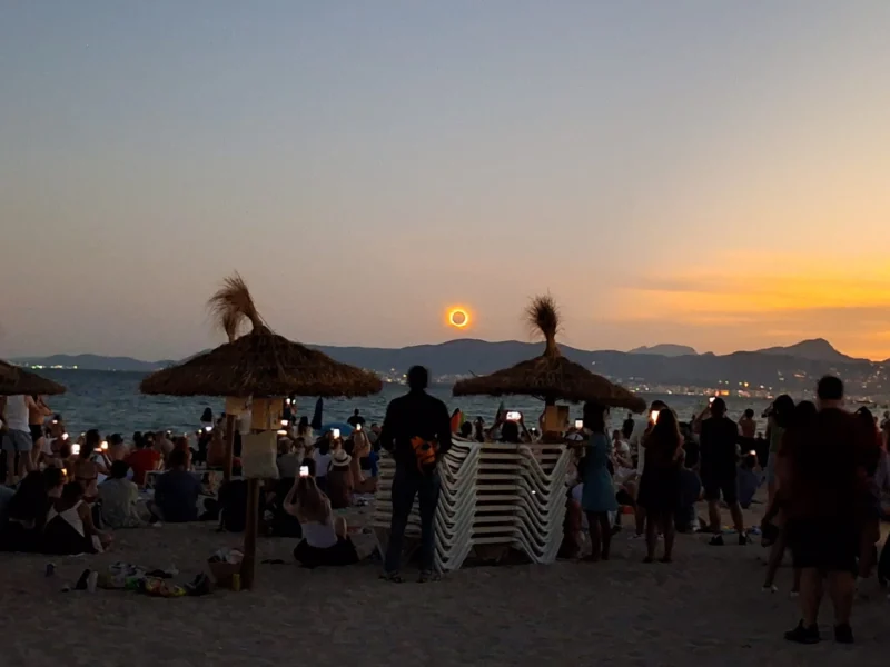 En menneskemængde samles på en strand ved solnedgang, hvor mange tager billeder af en solformørkelse over horisonten. Stråparasoller, stablede stole og det rolige hav kan ses under den dramatiske orange himmel.