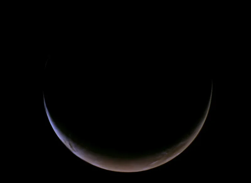 En tynd halvmåneformet planet, der for det meste ligger i skygge, med en svag blå og brun atmosfære, der kan ses langs dens solbelyste kant mod en sort rumbaggrund.
