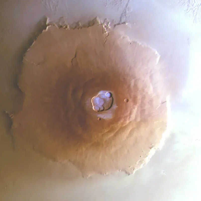 Luftfoto af Olympus Mons på Mars, der viser et stort, cirkelformet vulkansk bjerg med en central caldera og udstrålende skråninger, omgivet af en lys, støvet marsoverflade.