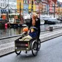 En kvinde kører på en ladcykel med to børn iført hjelme langs en kanal i en farverig by med både og historiske bygninger. Gaden er dekoreret med festlige guirlander.