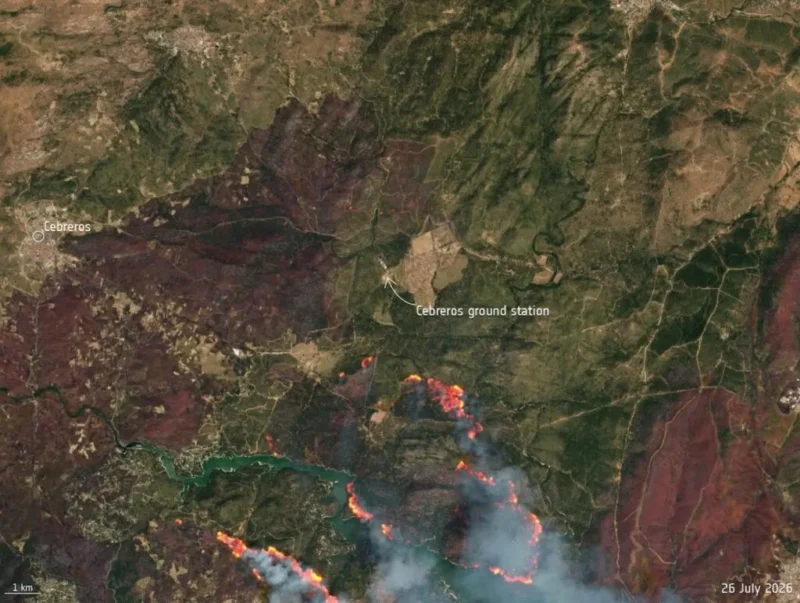 Satellitbillede, der viser Cerberos-jordstationen i nærheden af en skovbrand. I den nederste del ses orange flammer og røg, og der er angivet betegnelserne »Cerberos« og »Cerberos-jordstation«. Dato: 26. juli 2026. Målestok: 1 km.