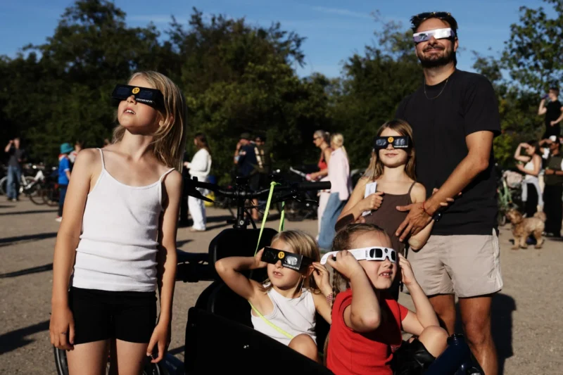 En gruppe børn og en mand har solformørkelsesbriller på og kigger op mod himlen i en park, hvor der er andre mennesker omkring dem, mens de nyder en solrig dag.