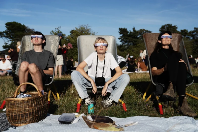Tre personer sidder på havestole på en græsplæne, iført solformørkelsesbriller og kigger op mod himlen. Foran dem ligger en picnickurv, en sodavandsdåse og et tæppe på jorden. I baggrunden samles andre mennesker.