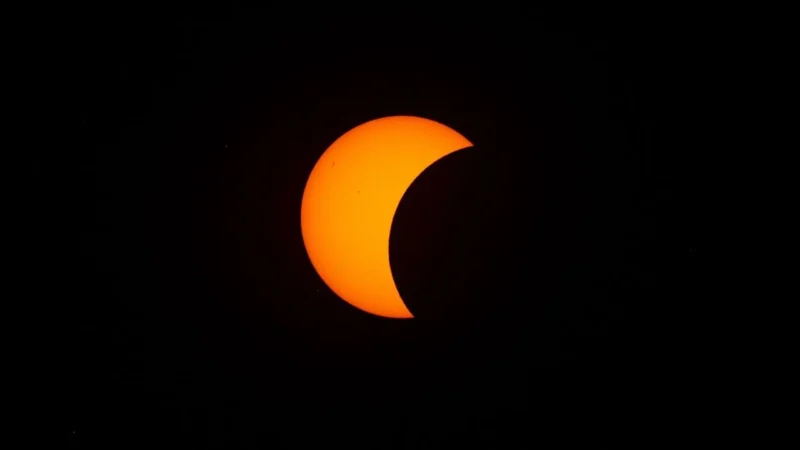 En lys orange delvis solformørkelse ses på en helt sort baggrund, hvor månen dækker en stor del af solen og danner en halvmåneform.
