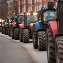 En lang række traktorer i forskellige farver er parkeret langs en bygade med bygninger i baggrunden og en cyklist, der kører forbi i venstre side af billedet.