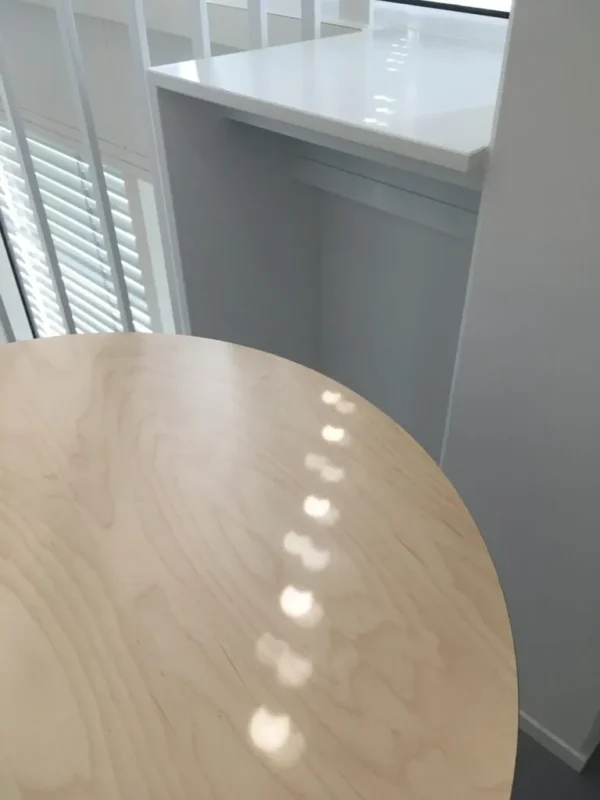 Et rundt bord af lyst træ med solpletter står tæt ved et hvidt gelænder og en lille, hvid hylde ved et vindue med persienner.