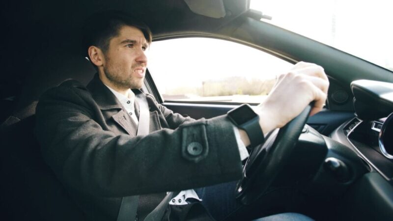 Man driving a car, wearing a seatbelt and a smartwatch, gripping the steering wheel with a focused expression.
