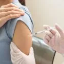 En sundhedsperson iført handsker giver en vaccineindsprøjtning i en persons overarm. Personen er iført en kortærmet skjorte og holder ærmet op for at modtage indsprøjtningen.