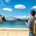 En mand med en rygsæk står på en sandstrand og kigger over vandet på Sydney Opera House og Sydney Harbour Bridge under en blå himmel med spredte skyer.
