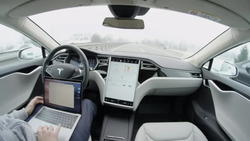 Et billede fra kabinen i en Tesla-bil, der kører på motorvejen i autopilot-tilstand. En person på førersædet bruger en bærbar computer med hænderne væk fra rattet, og den store berøringsskærm viser kørselsdata.