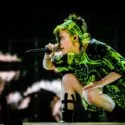 Billie Eilish er iført et grønmønstret outfit og pandebånd med sort tape på benet, og hun optræder under stærkt scenelys.