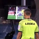 En fodbolddommer iført en neongul LEGEA-trøje gennemgår en situation på en videomonitor under en kamp og bruger Video Assistant Referee (VAR)-systemet til at træffe en afgørelse.