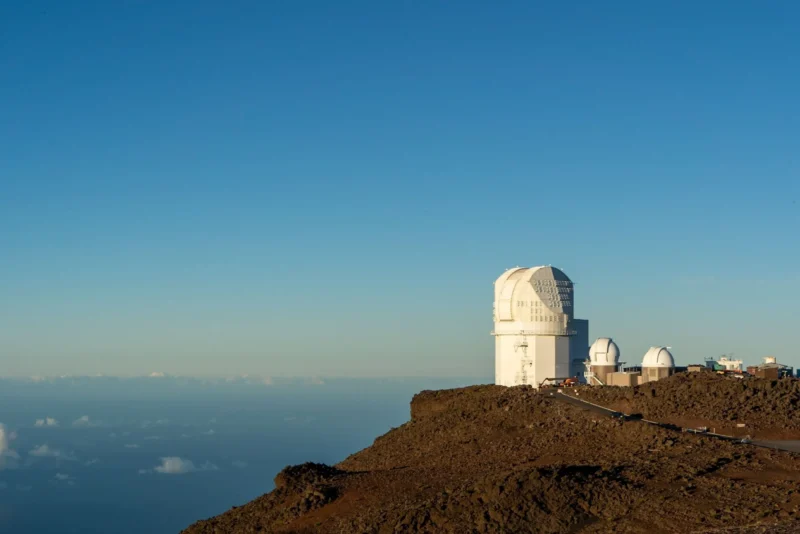 En stor hvid observatoriekuppel og to mindre kupler ligger på en klippefyldt bjergtop under en klar blå himmel, med udsigt over skyerne og havet i det fjerne.