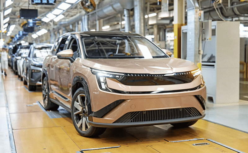 Front view of a pink Skoda SUV on an automotive assembly line in a factory setting, other cars and workers blurred in the background.