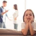 Pige sidder på kanten af sofa, mens far og mor skændes i baggrunden.