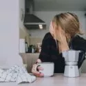 En person med rodet hår sidder ved et køkkenbord, holder et kaffekrus og dækker sit ansigt med den ene hånd. På bordet står en mokkakande og et ternet håndklæde, og i baggrunden ses et køkken.
