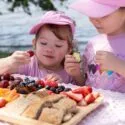 To unge piger i lyserøde skjorter og huer sidder ved et picnicbord ved vandet og nyder et fad med frisk frugt, grøntsager og brød på en solrig dag.