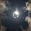 En solformørkelse med solen delvist dækket af månen, hvilket skaber en lysende korona og diamantringseffekt, omgivet af en mørk himmel og tynde, oplyste skyer.