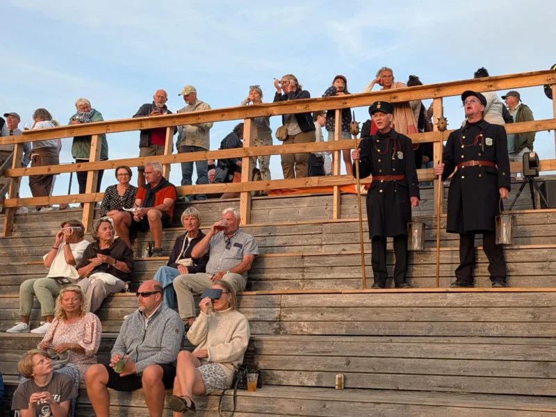 Folk sidder og står på trætribuner udendørs. To mænd i mørke uniformer står med stave i hænderne. Solen kaster et varmt lys, og himlen er klar. De fleste kigger på noget uden for billedet.