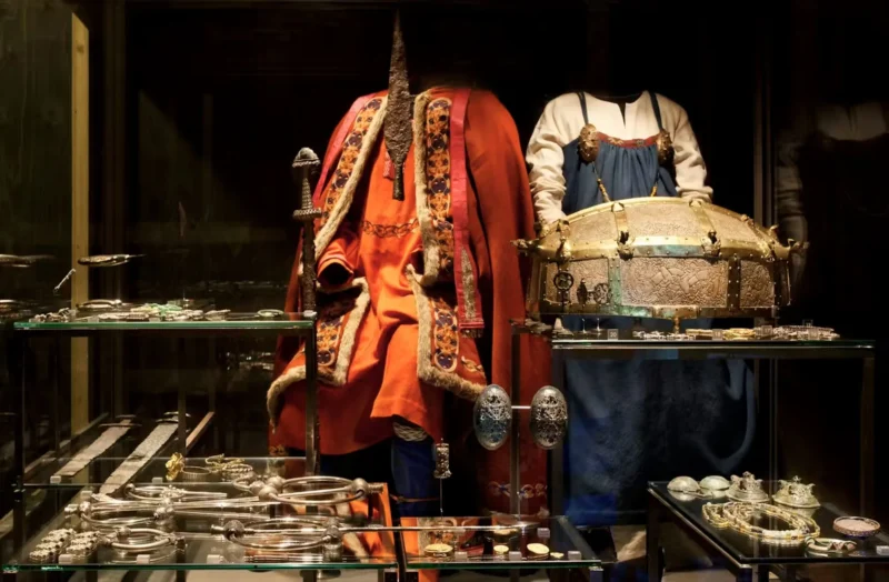 To udstillingsdukker viser middelalderligt tøj og udsmykkede smykker i en museumsvitrine. Sølvarmbånd, brocher og en kunstfærdig kiste er udstillet bag glas, hvor man kan se de kostbare stoffer og det dekorative metalarbejde.