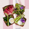 Fire åbne smørrebrød på mørkt brød, belagt med prosciutto, grønne blade, asparges, hårdkogte æg og spiselige blomster, anrettet på en baggrund, der ligner Union Jack-flagget i dæmpede farver.